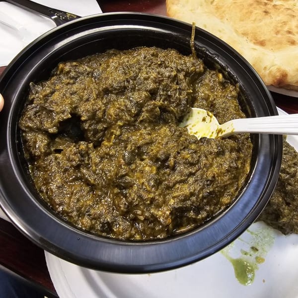 A black bowl of green spinach curry with a white spoon, served with naan bread.
